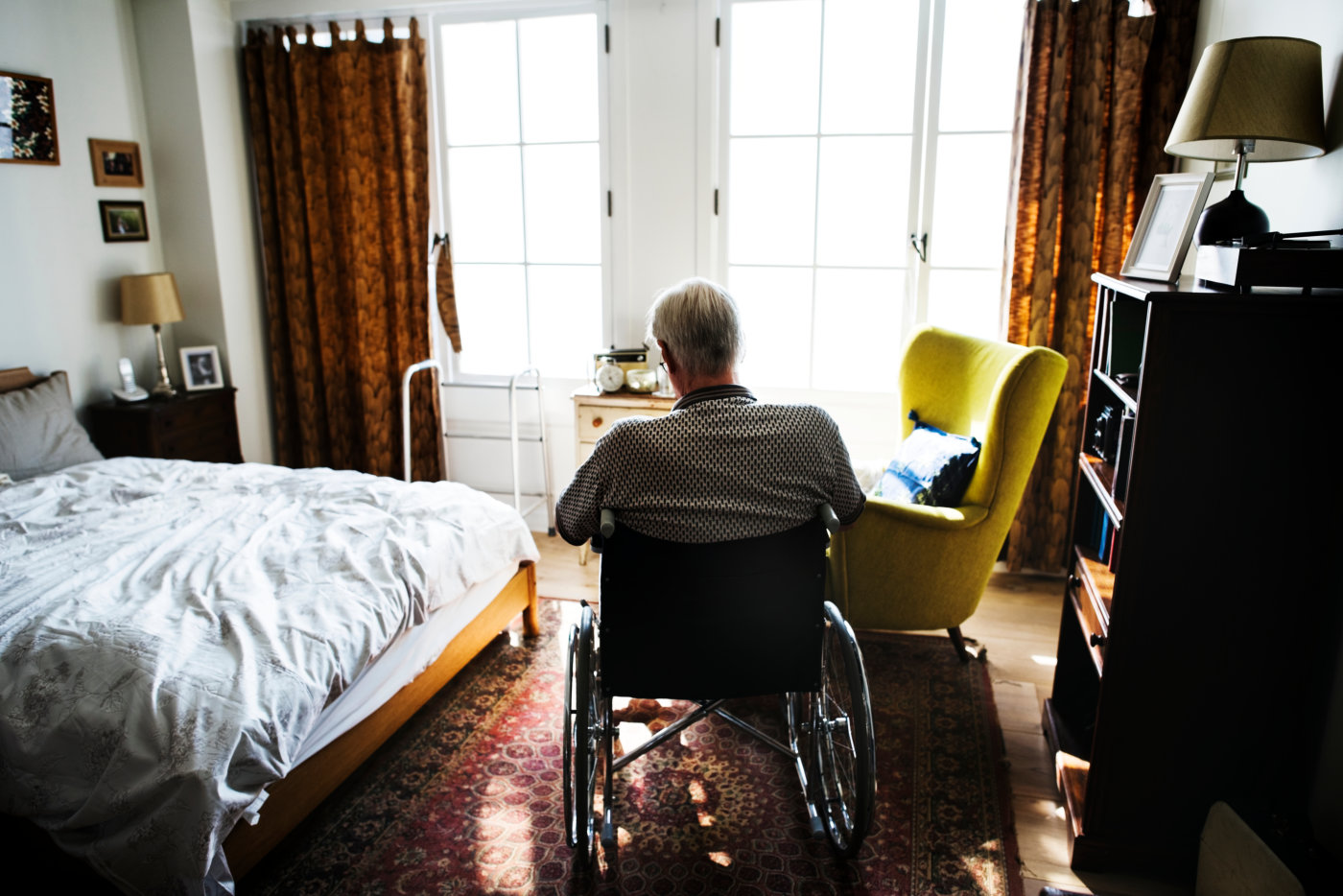 An elderly man in a wheelchair sits by a bed, reflecting a moment of calm amidst his Alzheimer's journey.