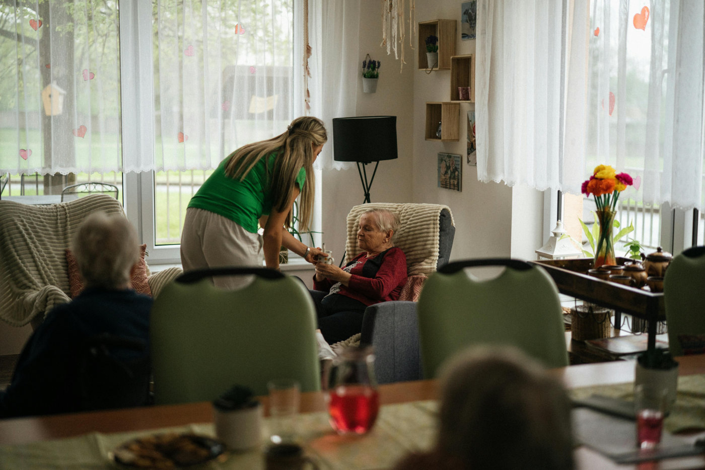 A caregiver assists an elderly woman seated in a cozy, sunlit room with large windows.