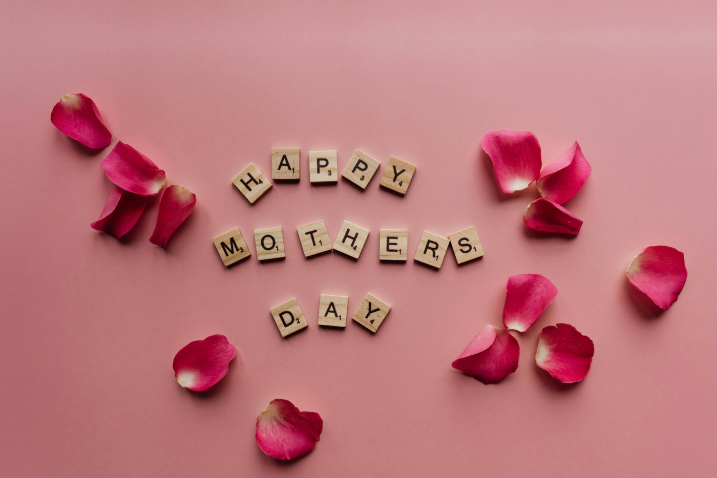 Happy Mother's Day spelled with Scrabble letters, surrounded by rose petals on a soft pink background.